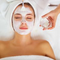 A woman lying with eyes closed while esthetician applies white facial mask at spa, towel-wrapped head