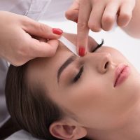 Close-up of a woman receiving eyebrow waxing while esthetician applies a wax strip to shape her brows.