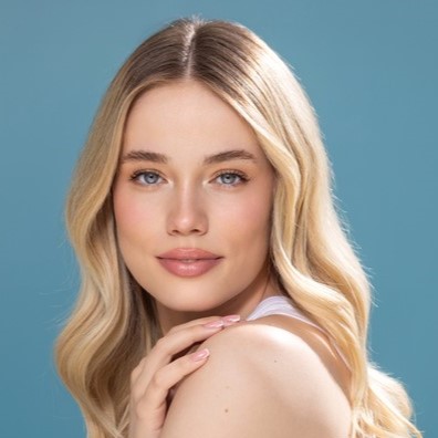 Close-up portrait of a young blonde woman with wavy hair, natural makeup and soft smile against a blue background.