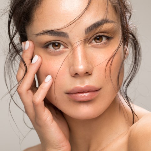 Beauty portrait close-up of a woman with clear skin, natural makeup, brown eyes, tousled hair, hand on cheek
