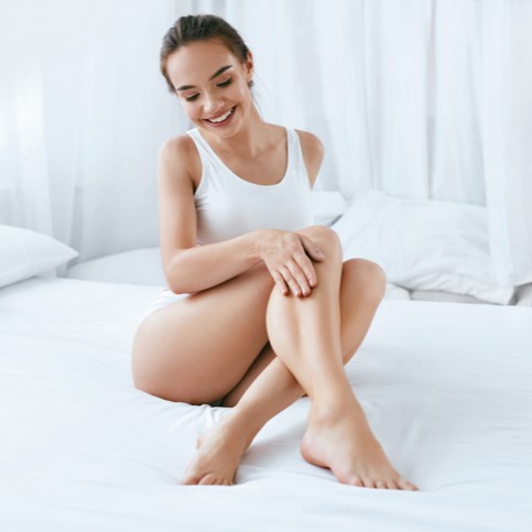 Smiling woman in white tank top sitting on bed, touching her legs, relaxed morning self-care scene.