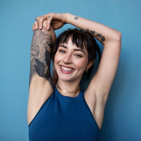 Smiling woman with tattooed arms wearing a blue tank top against a blue background